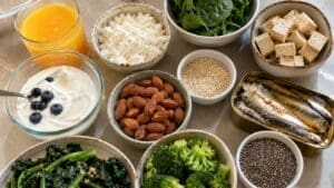 A variety of high calcium foods other than milk—including orange juice, spinach, tofu, cottage cheese, yogurt with blueberries, almonds, broccoli, sardines, kale, sesame seeds, and chia seeds—are arranged on a countertop.